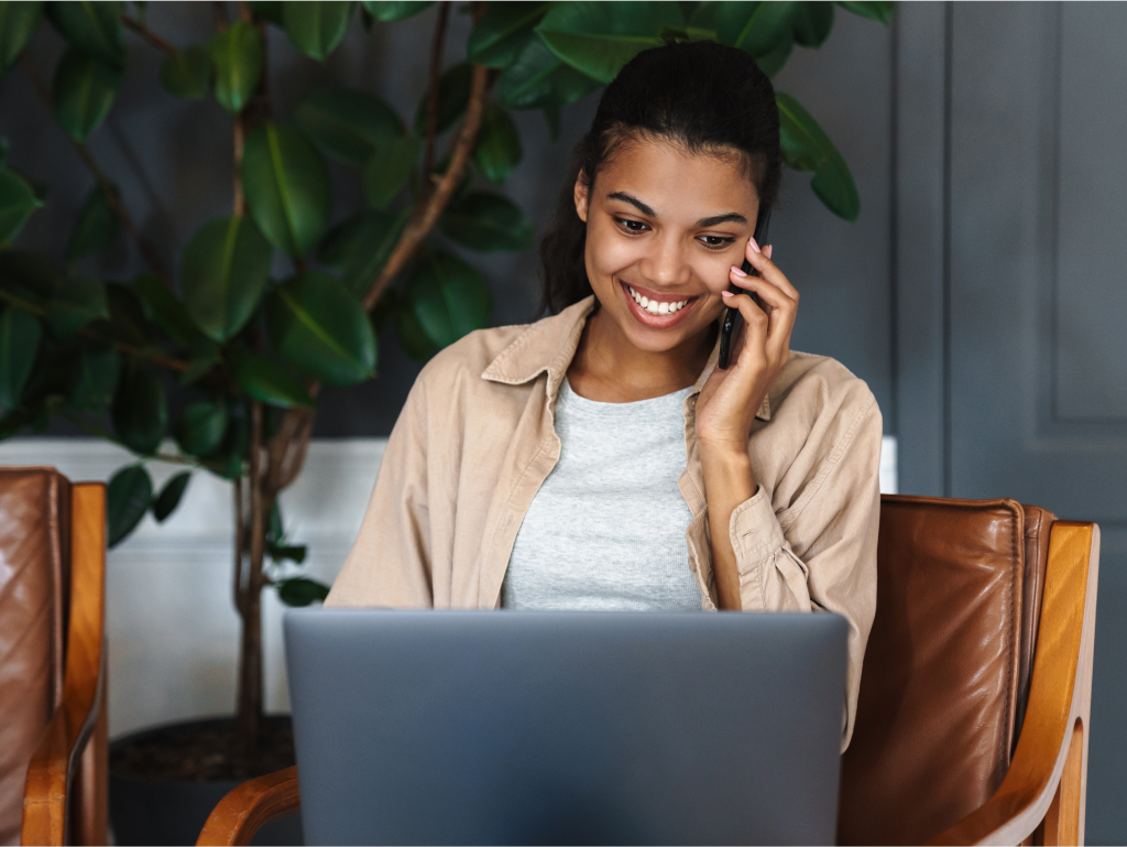 Smiling woman using her phone while working on a laptop, appearing happy and productive